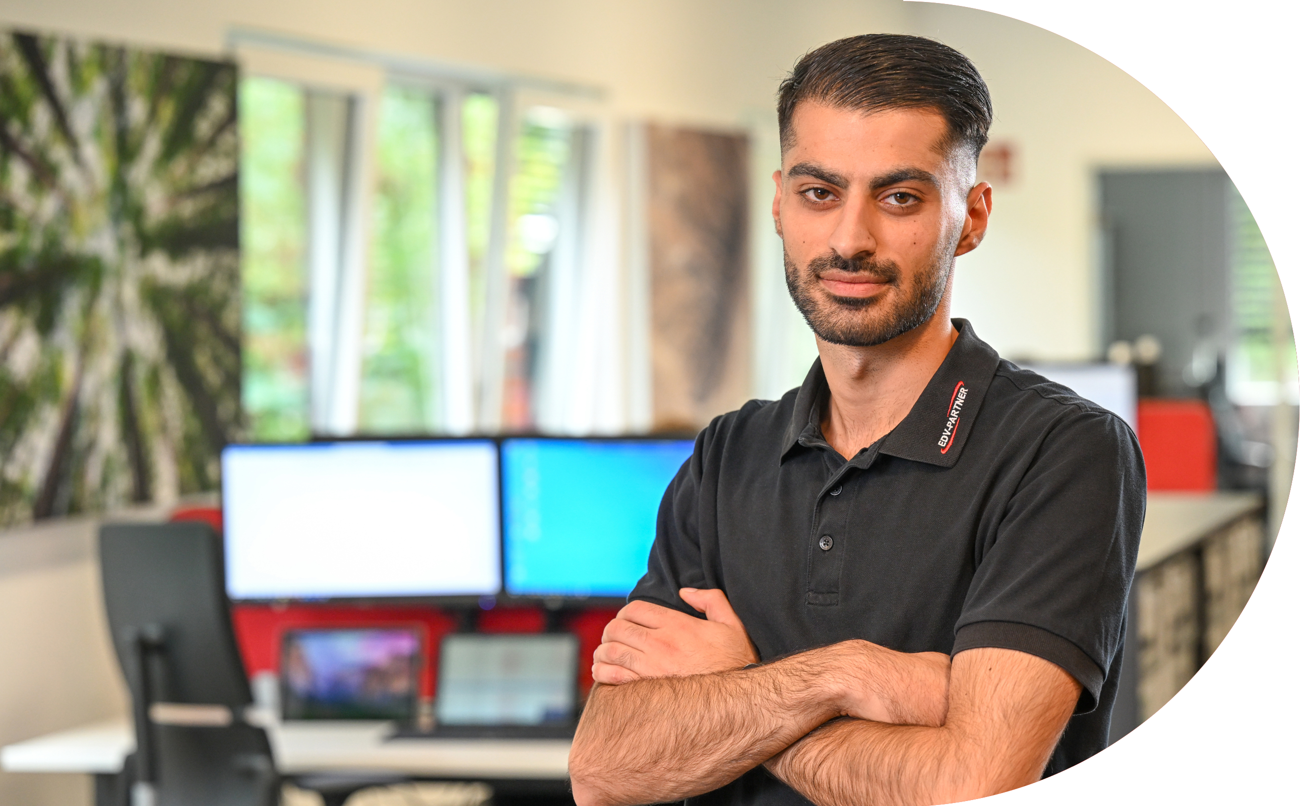Ein Mann mit schwarzen Haaren und Bart verschränkt seine Arme und schaut in die Kamera. Im Hintergrund sind mehrere eingeschaltete Bildschirme zu sehen