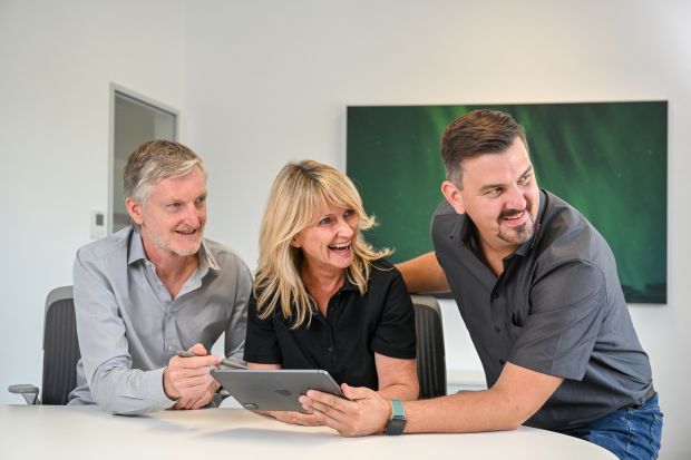 Drei lachende Personen sitzen gemeinsam am Tisch und schauen auf ein Tablet.